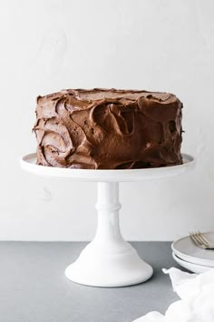 a chocolate frosted cake sitting on top of a white cake stand next to a plate