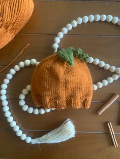 an orange knitted pumpkin and white beaded necklace on a wooden table with two knitting needles