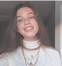 a woman with long brown hair wearing a white sweater and gold necklaces smiling at the camera