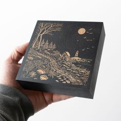 a person holding up a black box with gold foil on the lid and landscape painted on it