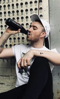 a man sitting on the ground drinking from a black bottle with his eyes closed while wearing a baseball cap