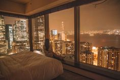 a person sitting on a bed in front of a window overlooking the city at night