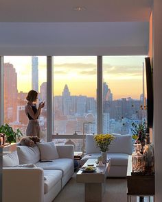 a woman standing in a living room next to a large window looking out at the city