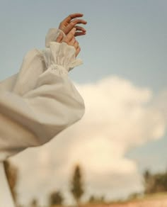 a woman wearing a white dress and holding her hand up to the sky with clouds in the background