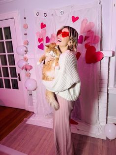 a woman holding a cat in her arms while standing next to a wall with hearts on it