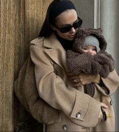 a woman in a trench coat holding a baby wearing a teddy bear hat and scarf