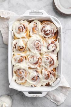 a white dish filled with cinnamon rolls on top of a marble counter next to a cup of coffee