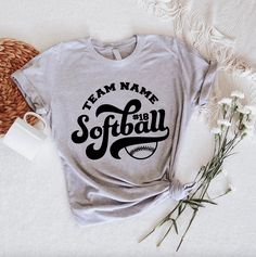 a t - shirt that says team name softball on it next to flowers and a cup