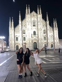 three girls posing in front of a cathedral at night with their arms up and one girl holding her hand up