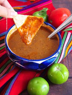 a hand holding a piece of bread over a bowl of salsa with apples and tomatoes in the background