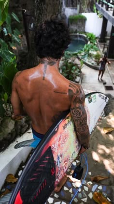 a man holding a surfboard while standing next to a tree and building with graffiti on it