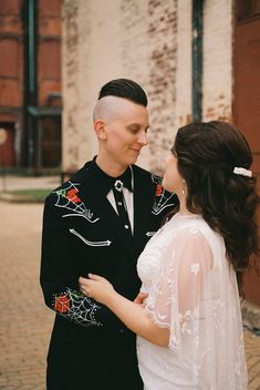 a man and woman standing next to each other in front of a brick building with red flowers on it