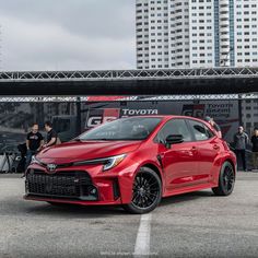 a red toyota car parked in front of some tall buildings with people standing around it