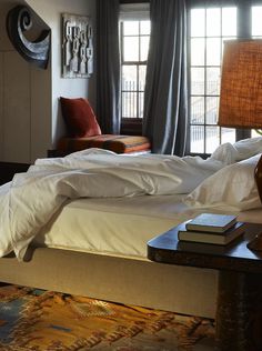 a bed with white sheets and pillows next to a lamp on a table in front of a window