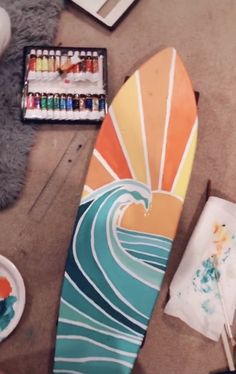 a surfboard sitting on top of a floor next to some paint and watercolors