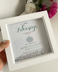 a person holding up a white frame with some flowers in the background and an arabic writing on it