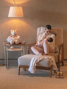 a woman sitting in a chair holding a baby next to a teddy bear and lamp