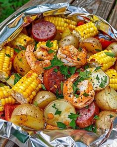 an aluminum foil container filled with shrimp, potatoes and corn on the cob covered in herbs