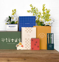 several notebooks, pens and flowers on a wooden shelf next to a brick wall