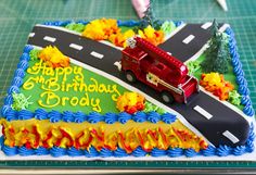 a birthday cake with a firetruck on the road and flowers in the background