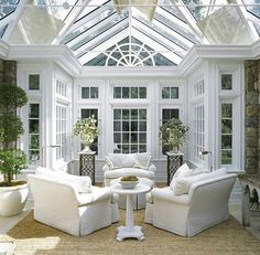 a living room filled with white furniture under a glass roof covered in windows and potted plants