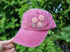 a pink hat with daisies embroidered on it in front of some green trees and bushes