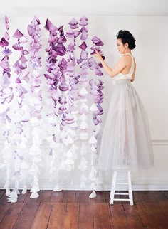 a woman standing on a ladder in front of a wall with purple paper flowers hanging from it