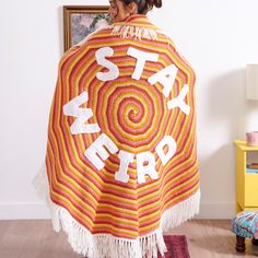 a woman is holding up a crocheted shawl with the words stay weird written on it