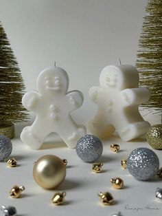 two white candles with silver and gold decorations next to small christmas trees on a table