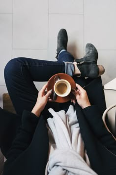 a person sitting on a couch holding a coffee cup and saucer with their feet up