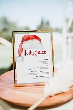 a sign with a santa hat on it sitting on top of a wooden table next to a white feather