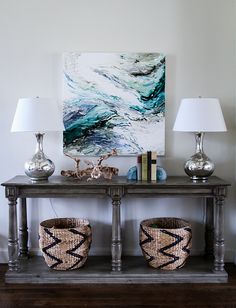 a table with two baskets on top of it next to lamps and a painting in the background