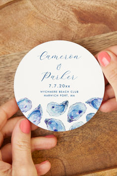 two hands holding up a white and blue round wedding sticker on a wooden table