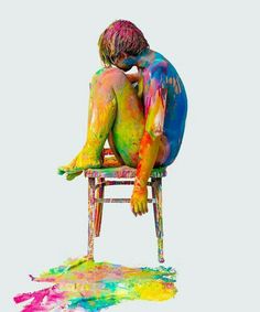 a man sitting on top of a wooden chair covered in colored paint and holding his head to his chest