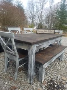 a wooden table with two chairs and a bench next to it on gravel covered ground