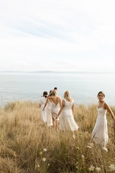 four women in white dresses walking through tall grass near the ocean on a sunny day