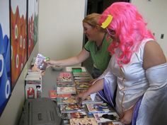 two women at a table with comic books and magazines on it, one woman has pink hair