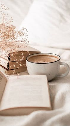 a cup of coffee sitting next to an open book on top of a white bed