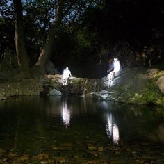 two people standing on rocks in the middle of a pond at night with lights reflecting off them