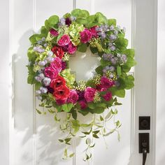 a wreath hanging on the front door of a house with flowers and greenery around it