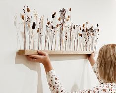 a woman holding up a piece of art with dried flowers on the wall behind her