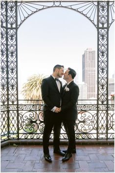 two men in tuxedos standing next to each other on a balcony with an arch