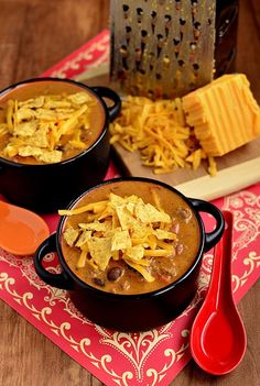 two black bowls filled with chili and cheese on top of a red place mat next to a grater