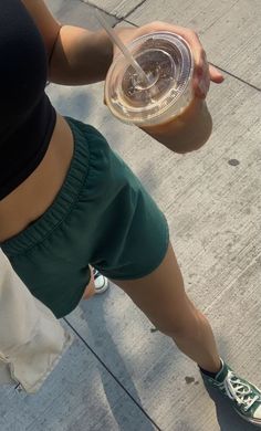 a woman is holding a drink in her right hand and wearing green shorts, while standing on the sidewalk