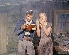 a man and woman standing next to each other in front of a stone wall holding an open book