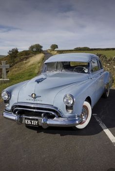 an old blue car is parked in a parking lot next to a stone wall and grass