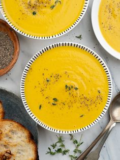 three bowls filled with soup next to bread and spoons on a white table top