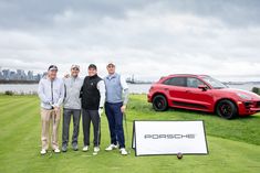 three men standing next to a red porsche cayenne on a golf course with a city in the background