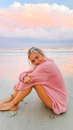 a woman sitting on top of a sandy beach under a pink sky with clouds in the background