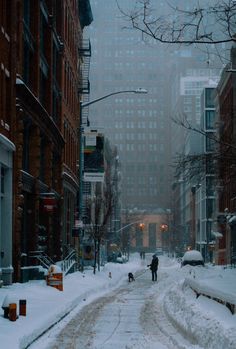 a man walking his dog down a snowy street in the city with tall buildings on either side